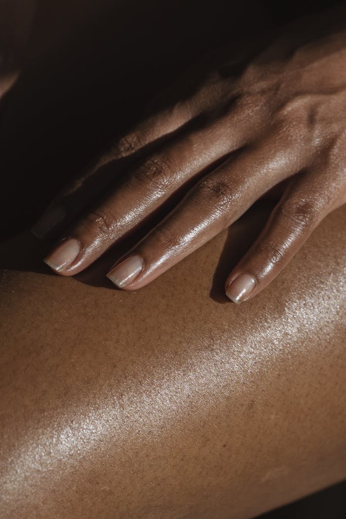 Crop anonymous African American female touching bare leg with hand while sitting on black background in room with bright sunlight